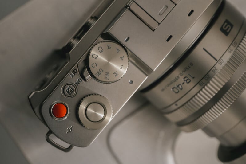 Close-up of a silver digital camera showing the top controls, including a silver mode dial, a red shutter button, and a lens labeled 18-40mm. The camera appears to be placed on a light surface.
