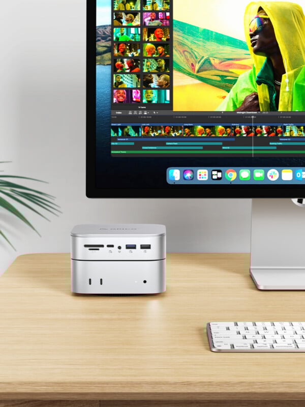 A modern desktop setup with a computer monitor displaying video editing software, a compact silver computer, a wireless keyboard, and a plant on a light wooden desk.