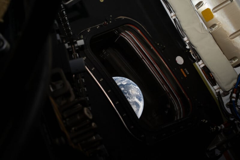 View of Earth through a window from inside a spacecraft, with part of the spacecraft’s interior and machinery visible around the edges of the window.