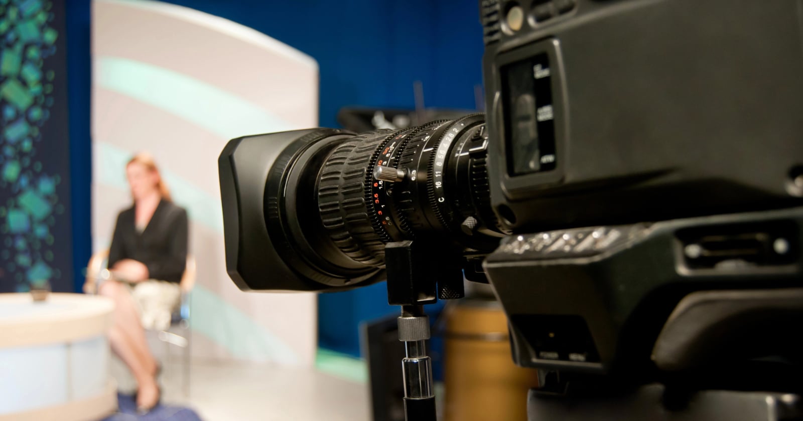 A close-up of a professional video camera focused on a blurred person sitting on a set, likely during a TV interview or news broadcast, with studio lights and a colorful backdrop.
