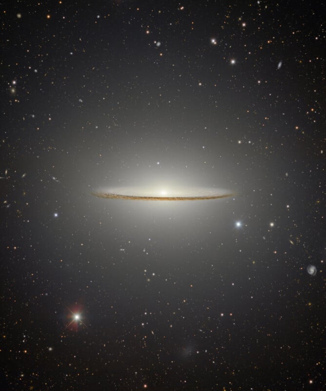 A glowing spiral galaxy, known as the Sombrero Galaxy, appears edge-on with a bright central bulge and a dark dust lane, surrounded by numerous distant stars and galaxies in deep space.
