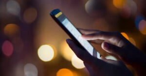 A close-up of a person's hand using a smartphone, with fingers touching the screen. The background is blurred with warm, colorful bokeh lights.
