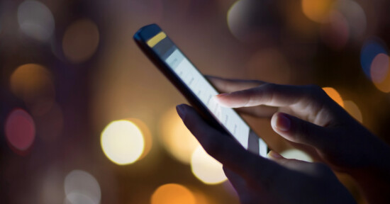 A close-up of a person's hand using a smartphone, with fingers touching the screen. The background is blurred with warm, colorful bokeh lights.