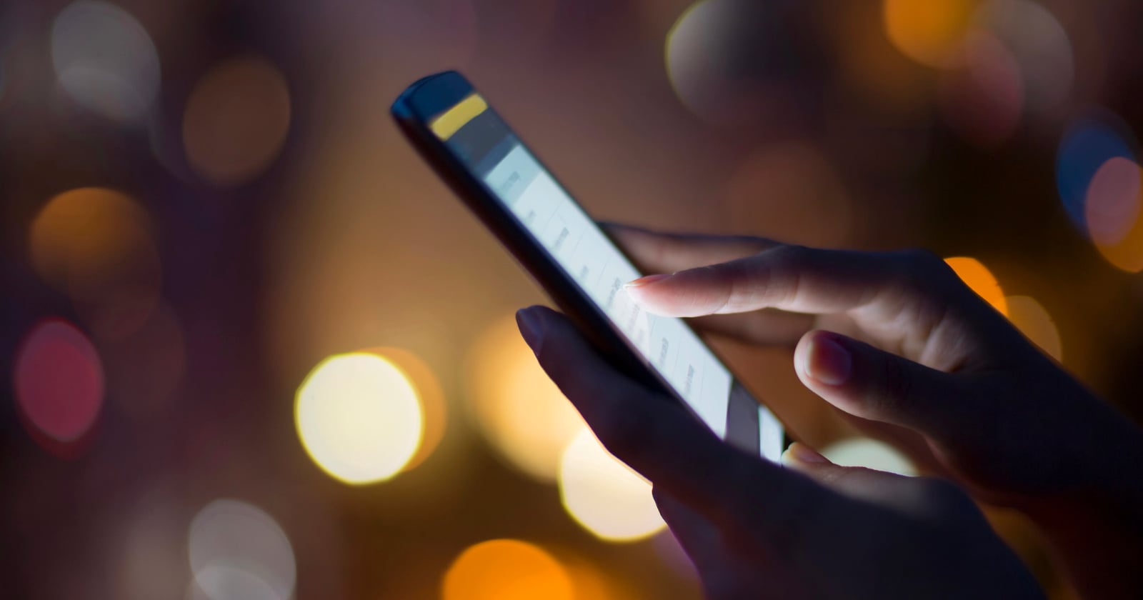 A close-up of a person's hand using a smartphone, with fingers touching the screen. The background is blurred with warm, colorful bokeh lights.