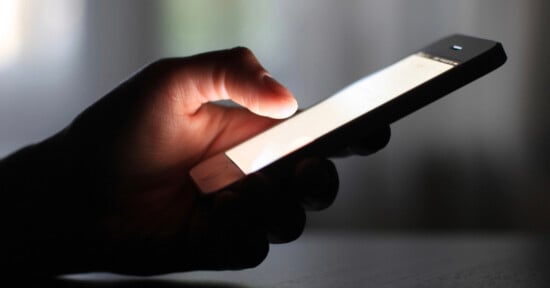 A close-up of a hand holding and using a smartphone in a dimly lit environment, with the phone screen brightly illuminated.