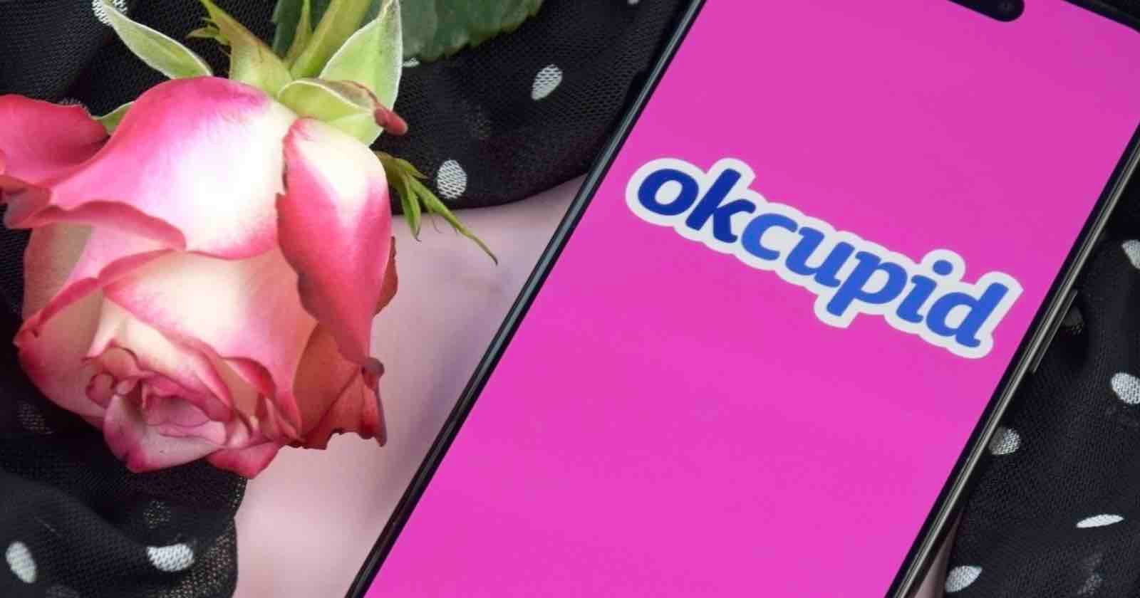 A smartphone with a pink screen displays the OkCupid logo. Next to the phone is a pink rose and a black fabric with white polka dots.