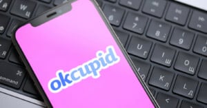 A smartphone displaying the OkCupid logo on a pink screen rests on a black computer keyboard with both English and Russian characters.
