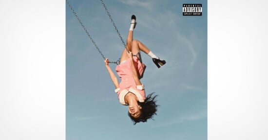 A young woman in a pink dress swings upside down against a clear blue sky, her hair hanging down and her mouth open as if yelling. She wears white socks and black shoes. The image has a parental advisory label in the corner.