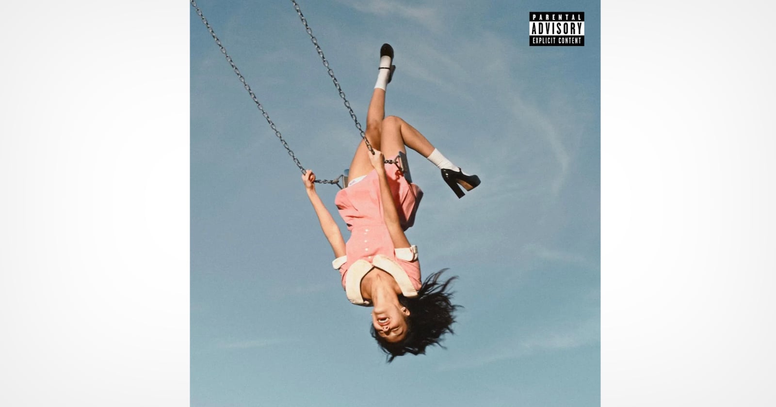 A young woman in a pink dress swings upside down against a clear blue sky, her hair hanging down and her mouth open as if yelling. She wears white socks and black shoes. The image has a parental advisory label in the corner.