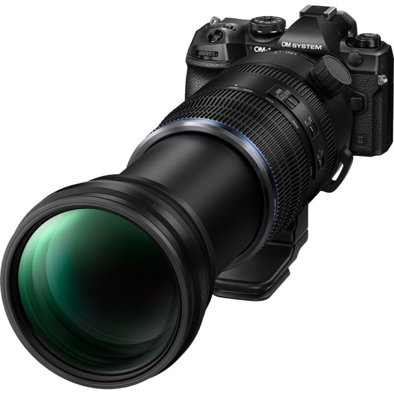 A black digital camera with a large, long telephoto lens attached, featuring textured grip rings and a reflective green front element. The camera body displays various buttons, dials, and the "OM SYSTEM" branding.