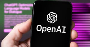 A hand holds a smartphone displaying the OpenAI logo and text on the screen. The blurred background features a computer monitor with purple and green hues and indistinct text.