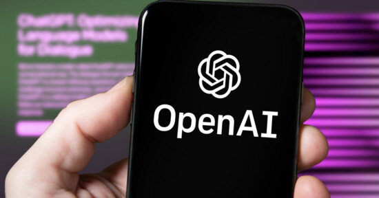 A hand holds a smartphone displaying the OpenAI logo and text on the screen. The blurred background features a computer monitor with purple and green hues and indistinct text.