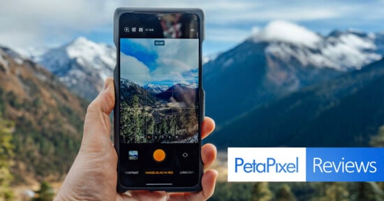 A hand holding a smartphone that is taking a photo of a scenic mountain landscape with snow-covered peaks and a forest, with the Petapixel Reviews logo at the bottom right.