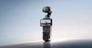 A compact handheld camera with a built-in gimbal stands upright. Its screen displays a landscape with mountains at sunset. The device has several buttons and a joystick on the front. The background is softly lit and minimalistic.