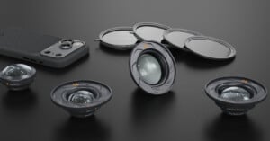 Four camera lenses, three round lens filters, and a smartphone with a textured back are arranged on a dark surface, showcasing photography accessories in a neat layout.