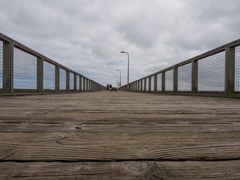 A low-angle view of a wooden pier with metal railings on both sides, stretching into the distance under a cloudy sky. A few people are visible far away near the end of the pier.