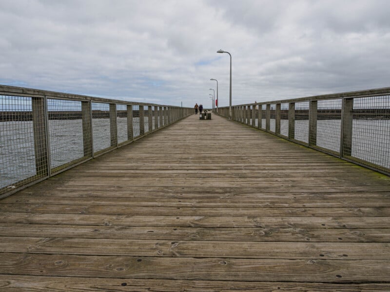 A wooden pier with metal railings extends over the water under a cloudy sky. Two people walk in the distance near a lamp post at the end of the pier.