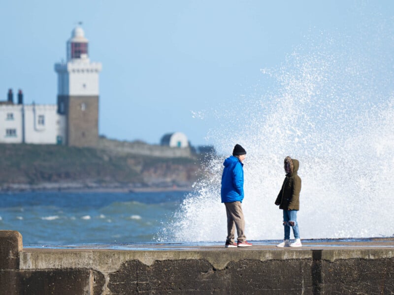 Two children stand on a seaside pier, talking, as a large wave splashes behind them. In the background, a lighthouse and building are visible across the water under a clear blue sky.