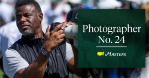 A man holding a camera with a large lens stands outdoors. Beside him, a green sign reads "Photographer No. 24 Masters" with the Masters golf tournament logo.