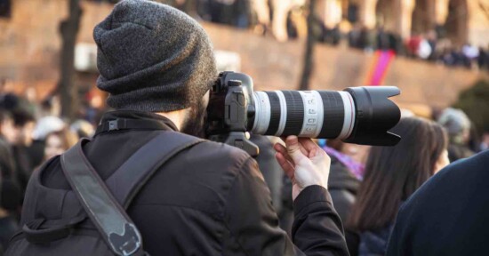A person wearing a beanie and backpack holds a professional camera with a large telephoto lens, photographing a crowd of people outdoors.
