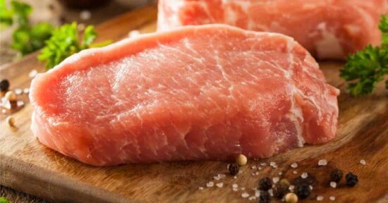 Raw pork chops on a wooden cutting board, surrounded by parsley, coarse salt, and whole peppercorns.