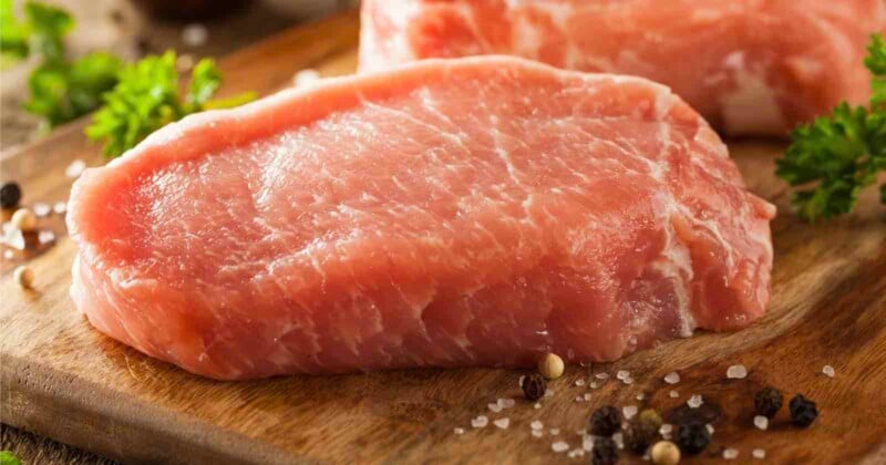 Raw pork chops on a wooden cutting board, surrounded by parsley, coarse salt, and whole peppercorns.