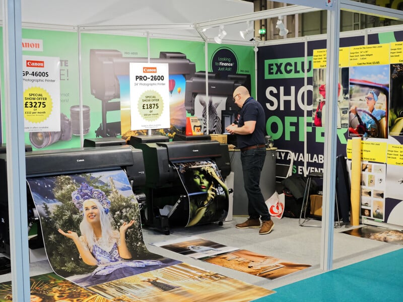 A man stands at a trade show booth featuring large Canon printers, with vibrant, oversized photo prints emerging from the machines. Promotional signs display special offers on the printer models.