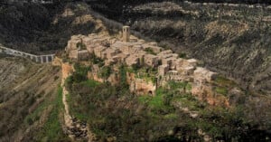 Aerial view of a small medieval village with stone buildings and a tall bell tower, perched atop a steep, rocky hill surrounded by green vegetation and deep valleys.