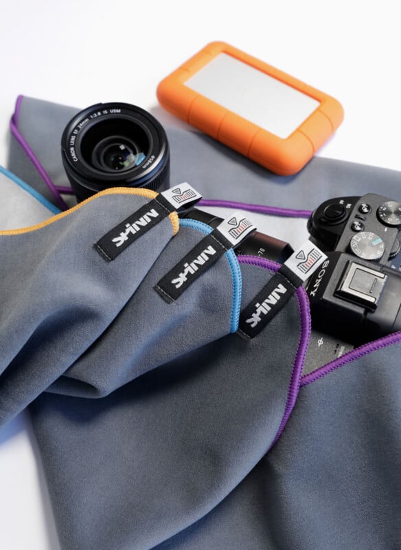A camera, lens, and orange portable hard drive rest on a gray microfiber cloth with colorful, stitched edges and visible "MINIF" brand tags.