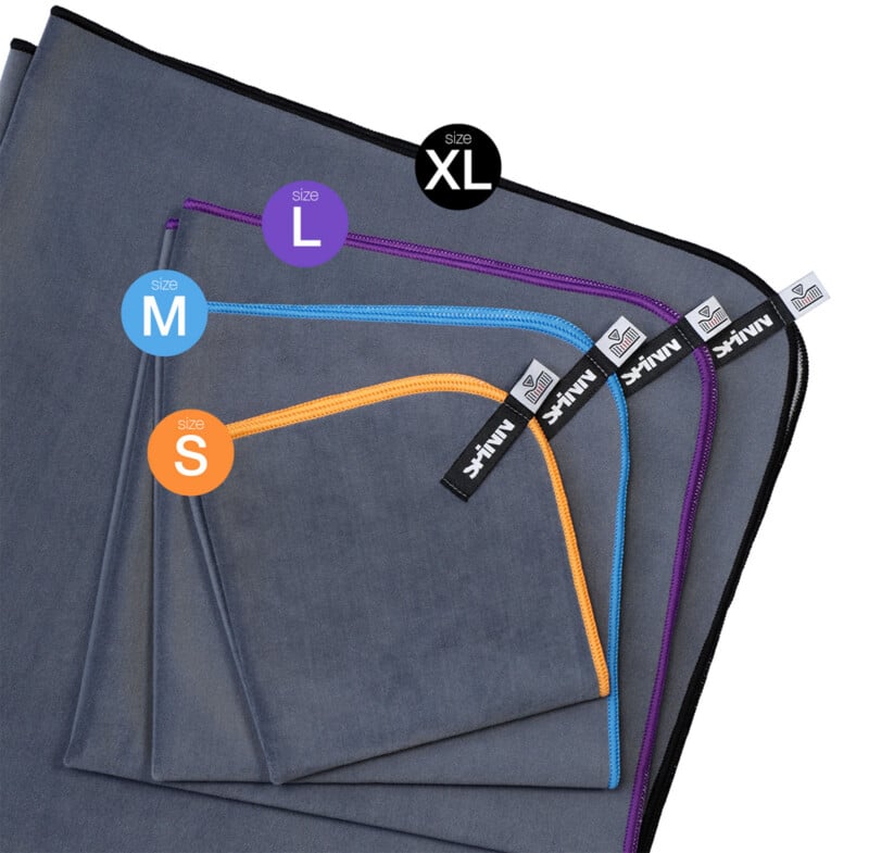 Four gray microfiber towels in sizes XL, L, M, and S are stacked, each with colored trim and labeled tags: black for XL, purple for L, blue for M, and orange for S.