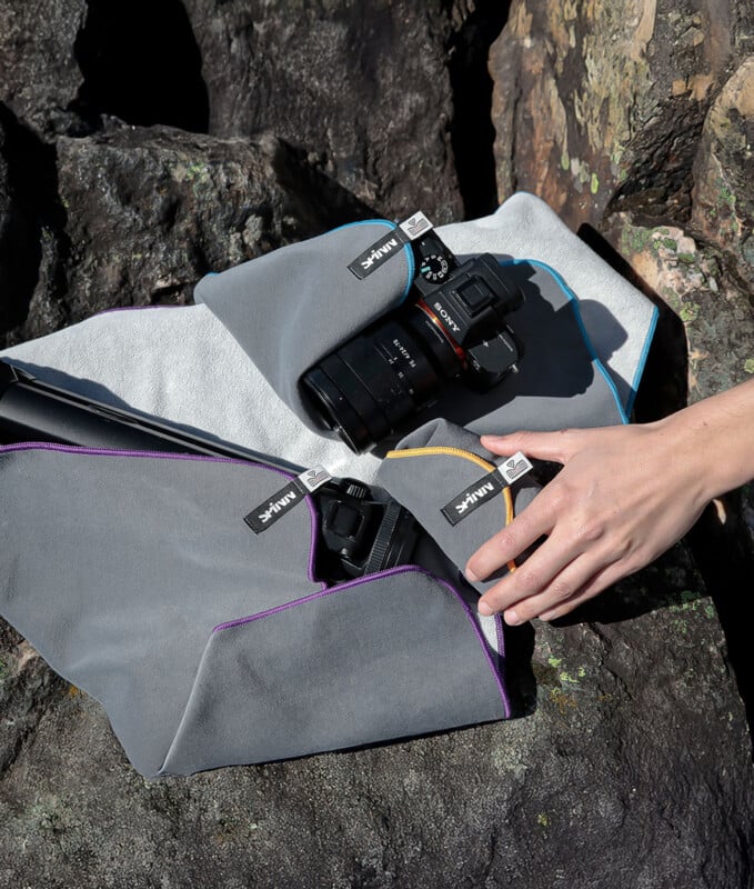 A hand wraps a camera and lens in gray padded cloths with colored trim, placed on rocky outdoor ground. The cloths have visible tags marked “Hama.”.