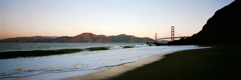 A panoramic view of a sandy beach at sunset with gentle waves, the Golden Gate Bridge in the distance, and hills in the background under a clear sky.