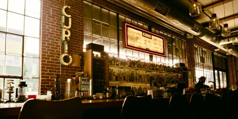 A dimly lit bar with high ceilings, exposed brick walls, shelves of liquor bottles, a glowing "CURIO" sign, and a large menu board above the bar. A person sits at the bar near large windows.