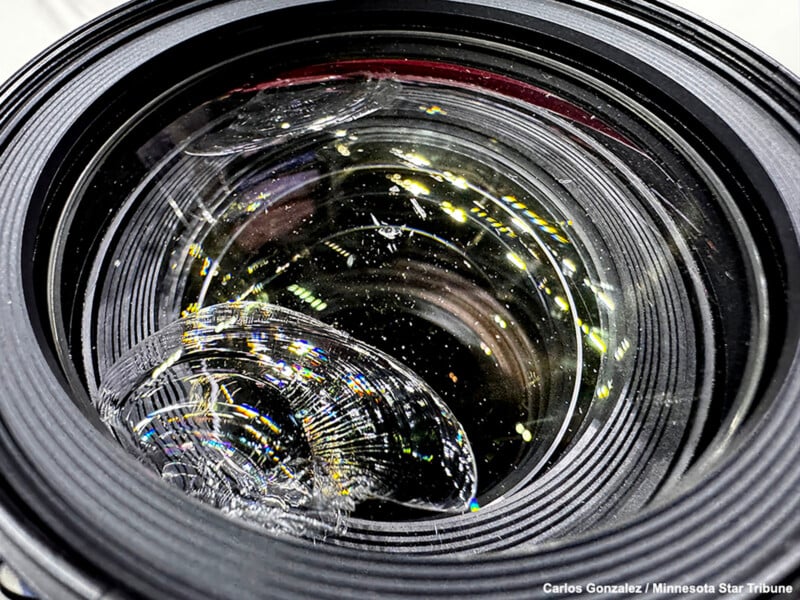 Close-up of a camera lens with a large, noticeable crack and shattered glass in the lower left area, surrounded by lens reflections and light specks.