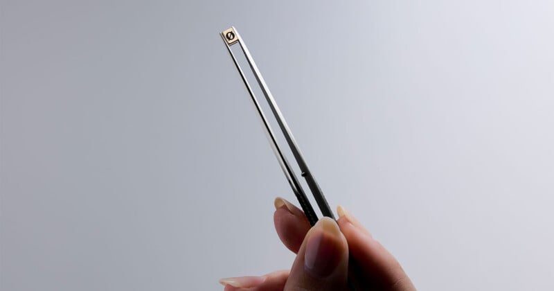 A hand with long fingernails holds a thin, rectangular, metallic object vertically against a plain, light gray background.