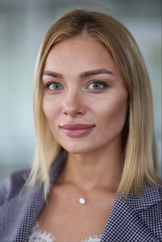 A woman with straight blonde hair, green eyes, and natural makeup wears a gray checkered blazer and a delicate necklace, smiling softly at the camera against a blurred background.