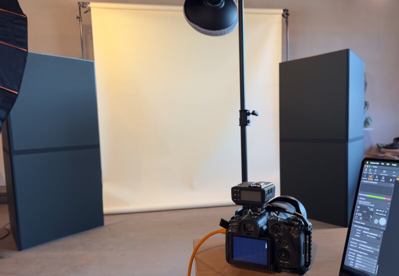 A photography studio setup with a digital camera on a tripod, lighting equipment, two large dark panels, a computer screen showing camera controls, and a beige backdrop.