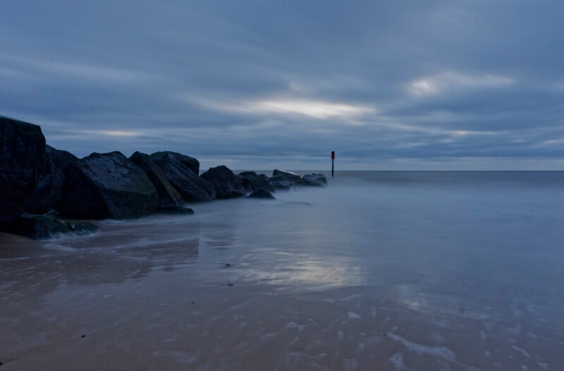 A rocky shoreline at dusk with calm waves washing over the sand, dark rocks extending into the sea, and a cloudy sky casting a blue-grey hue across the tranquil scene.