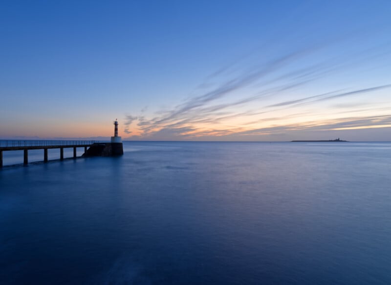 A tranquil seascape at sunset with a lighthouse on a pier extending into calm blue water. The sky is streaked with clouds, and an island is visible on the horizon under soft, fading light.