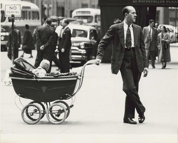 A man in a suit leans casually on a baby stroller while standing on a busy city sidewalk; pedestrians and cars fill the background, with a "No Parking" sign visible.
