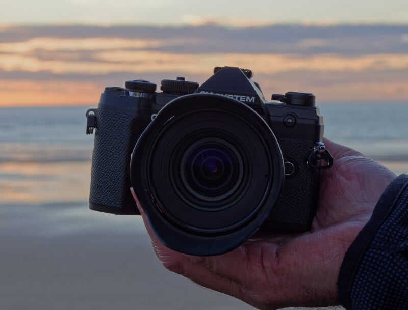 A close-up of a hand holding a black digital camera with a wide lens, against a blurred background of a beach and ocean at sunset.