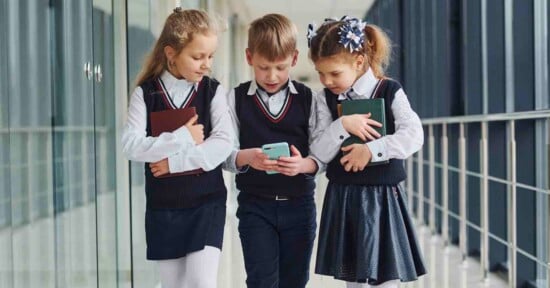 Three schoolchildren in uniforms walk down a hallway. The boy in the middle looks at a smartphone, while the two girls on either side hold books and look at the screen with interest.