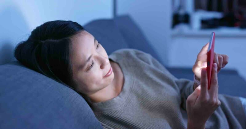 En una habitación con poca luz, una mujer está tumbada en un sofá gris, sonriendo y mirando su teléfono inteligente. La suave luz del teléfono inteligente le da en la cara.
