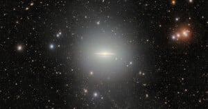 A bright, flat galaxy resembling a glowing disk is centered in a star-filled space, surrounded by numerous stars and distant galaxies against a dark sky.