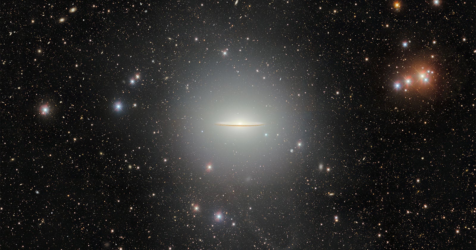 A bright, flat galaxy resembling a glowing disk is centered in a star-filled space, surrounded by numerous stars and distant galaxies against a dark sky.