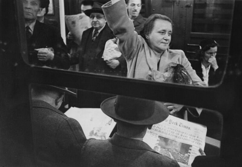 Black-and-white photo of people inside a subway car, some sitting and reading newspapers, while others stand holding onto straps. A woman stands in the center, arm raised, looking ahead with a calm expression.