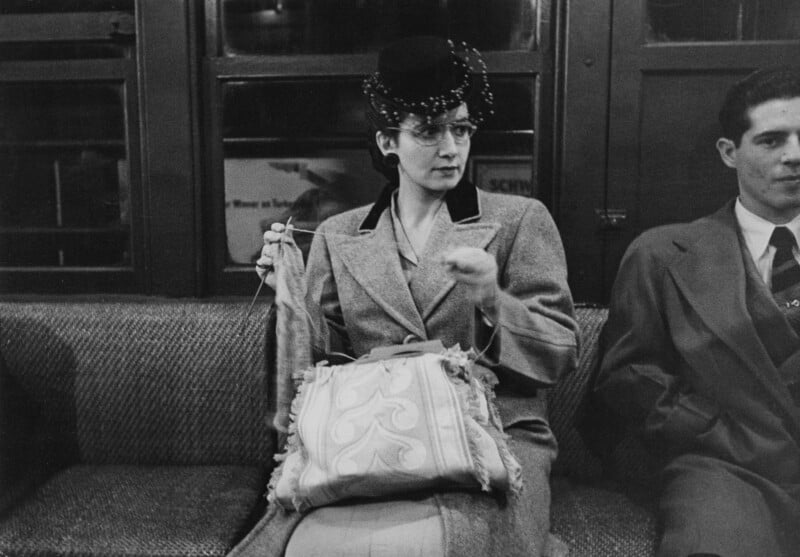 A woman in a hat and coat knits on a subway train, seated next to a man in a suit. She holds her knitting and looks to the side. The scene appears to be from an earlier era, possibly the mid-20th century.