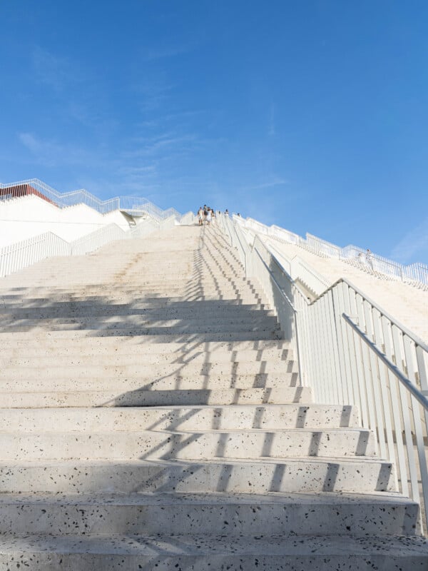Wide stone staircase with metal railings ascending steeply under a clear blue sky. Several people are visible at the top, with shadows cast by the railings onto the steps.