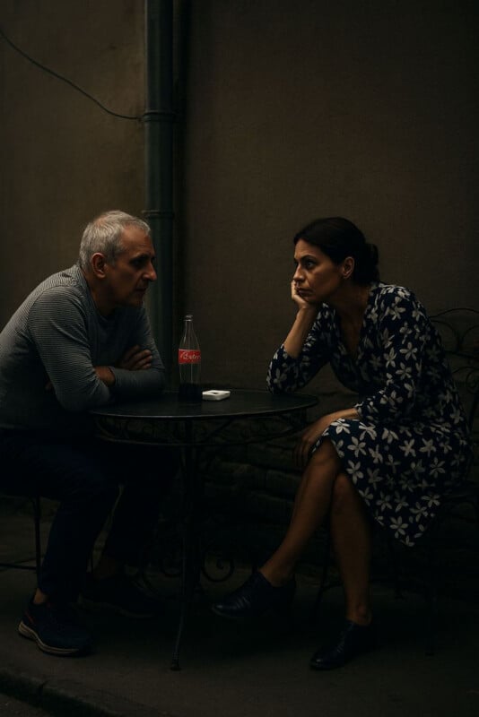A man and a woman sit at a small round table outdoors, engaged in a serious conversation. The woman rests her head on her hand, looking pensive. A glass bottle of Coca-Cola and a pack of cigarettes are on the table.