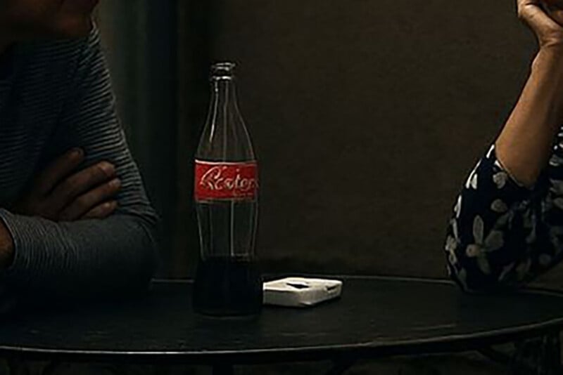 Two people sit across from each other at a round table. On the table are a glass bottle with a red label and a white rectangular object. Their faces are not visible, and the scene is dimly lit.